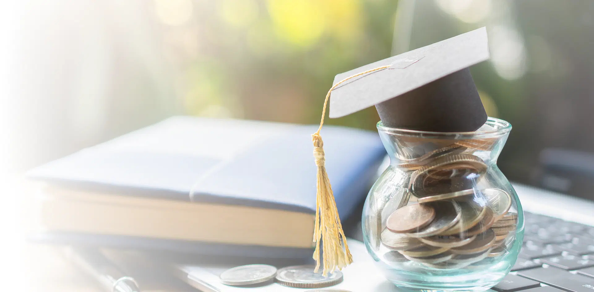 Graduation hat model inside a jar with money coins next to a book placed on a laptop.